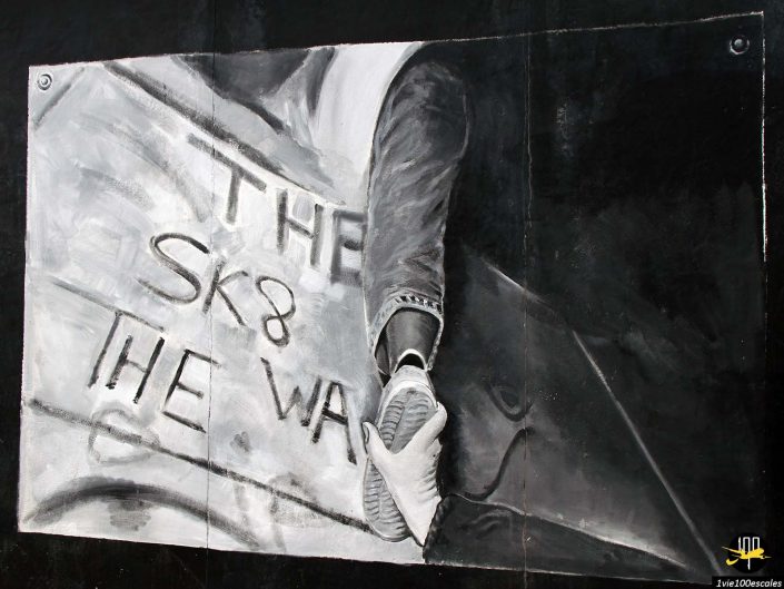 Image en noir et blanc d'une personne tenant un skateboard, avec une pancarte indiquant "THE SK8 THE WA", partiellement visible, prise à Berlin en Allemagne.