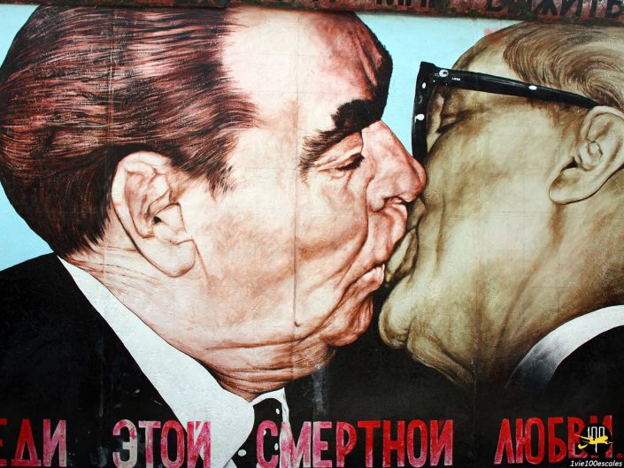 Une fresque murale à Berlin en Allemagne représente deux hommes s'embrassant. Les deux hommes portent des costumes et l’image est stylisée avec des couleurs vives. Le texte russe est visible au bas de la fresque.