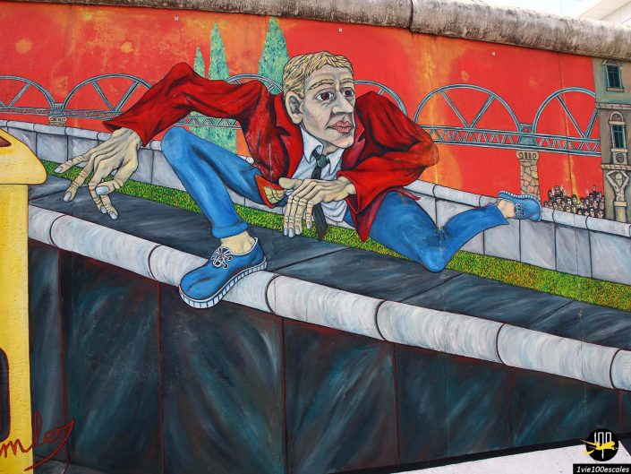 Une fresque murale colorée représente une personne vêtue d'une veste rouge et de chaussures bleues escaladant une barrière avec un pont et des bâtiments en arrière-plan, à Berlin en Allemagne.