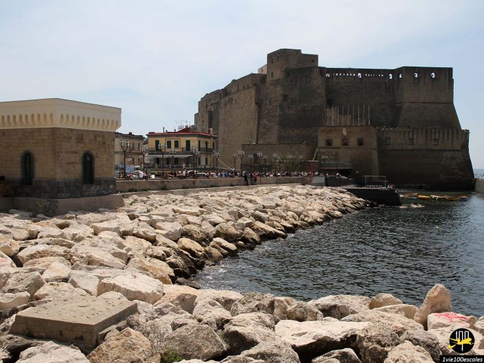 Château en bord de mer avec côte rocheuse, bâtiments et promenade au bord de l'eau à Naples en Italie. Les gens marchent le long de la promenade au bord de l’eau.