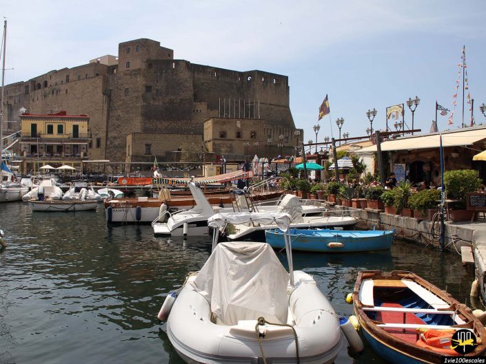Une marina avec divers bateaux amarrés devant une forteresse historique à Naples en Italie. Le quartier est animé avec des restaurants et des gens qui se promènent et profitent de l'atmosphère animée.