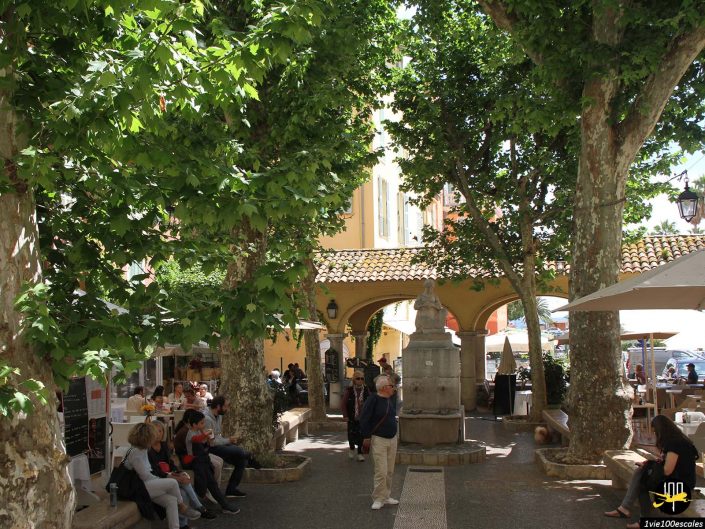 Des gens assis et marchant autour d'une place ombragée avec de grands arbres, une fontaine centrale et des bâtiments environnants avec des sièges de café à Menton en France.