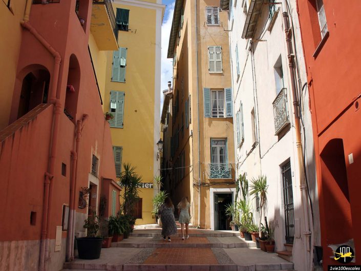 Une rue étroite flanquée de bâtiments colorés à plusieurs étages avec des volets verts et des balcons à Menton en France. Deux personnes s'éloignent au loin, des plantes en pot bordent la rue et des escaliers mènent à une porte cintrée sur la gauche.