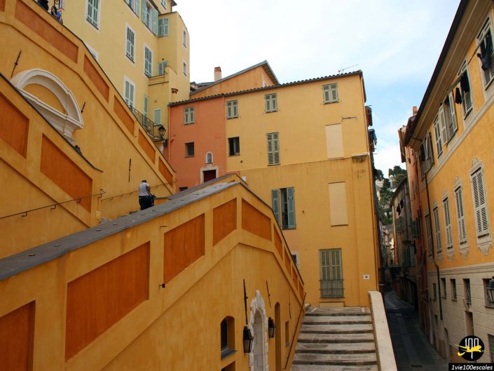 Une personne monte un large escalier entre des bâtiments colorés avec des volets à Menton en France ; le chemin diverge à gauche et à droite en haut.