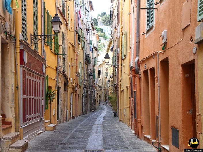 Une rue étroite et vide bordée de bâtiments colorés s'étend vers une colline au loin à Menton en France. Du linge est suspendu aux fenêtres et plusieurs lampadaires sont fixés aux murs.