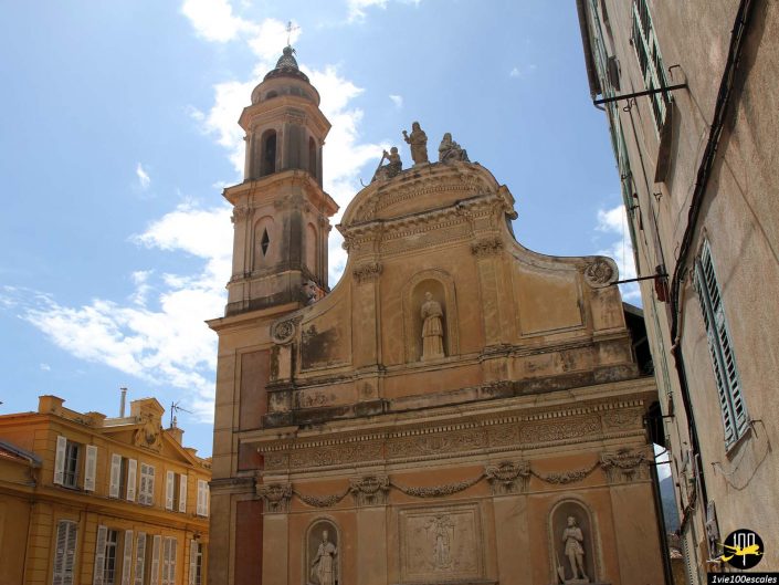 Une église historique avec un clocher se dresse sous un ciel bleu à Menton en France. La façade présente des sculptures et des statues détaillées, tandis que les bâtiments adjacents sont visibles sur la gauche.