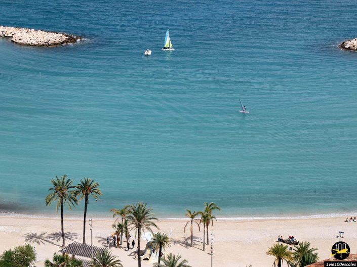 Vue aérienne d'une plage à Menton en France avec des palmiers épars, quelques personnes marchant le long du sable, et une eau turquoise calme où l'on aperçoit deux voiliers et une planche à voile.