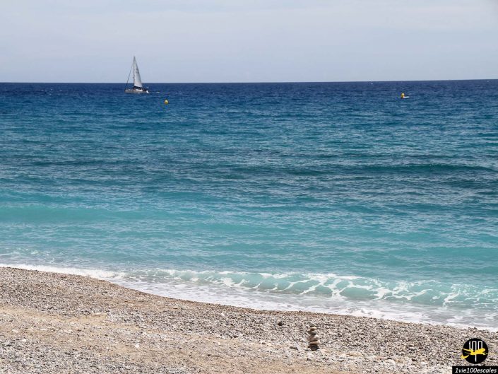 Un voilier flotte sur la mer bleue et calme près d'une plage de galets avec de petites pierres empilées au premier plan, à Menton en France.