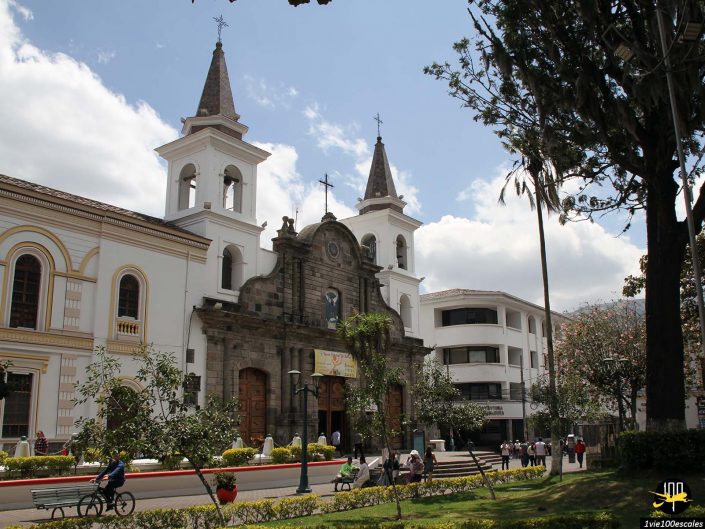 Une église historique avec deux clochers est vue depuis une place à Ibarra en Équateur, avec des gens assis sur des bancs et marchant. Des arbres et un ciel bleu avec des nuages sont en arrière-plan.
