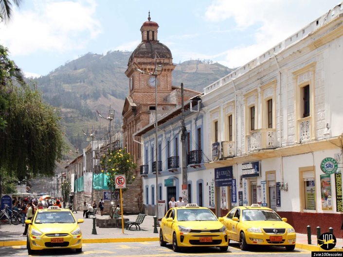 Scène de rue avec trois taxis jaunes garés devant des bâtiments historiques, dont une importante tour de l'horloge, sous un ciel clair avec des montagnes en arrière-plan à Ibarra en Équateur.