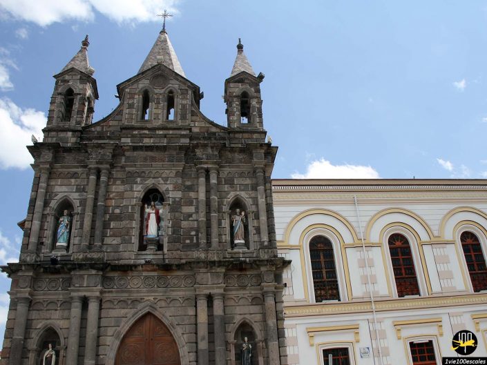 Façade d'église en pierre avec trois statues situées dans des niches cintrées, adjacente à un bâtiment blanc et or aux fenêtres cintrées, sous un ciel bleu avec quelques nuages, à Ibarra en Équateur.