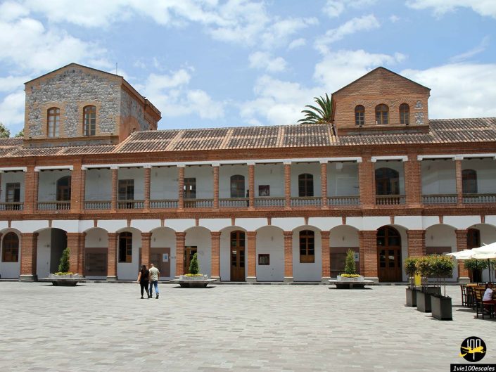 Un bâtiment historique avec une grande place ouverte devant se dresse gracieusement à Ibarra en Équateur. La structure présente une construction en brique et en pierre avec des fenêtres cintrées et des balustrades. Trois personnes se tiennent sur la place sous un ciel partiellement nuageux.
