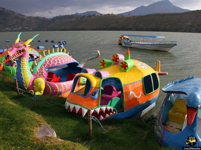 Des pédalos colorés sur le thème du dragon et du poisson sont garés au bord du lac, avec un plus grand bateau d'excursion flottant en arrière-plan. Un ciel nuageux et un paysage montagneux sont visibles au loin à Ibarra en Équateur.
