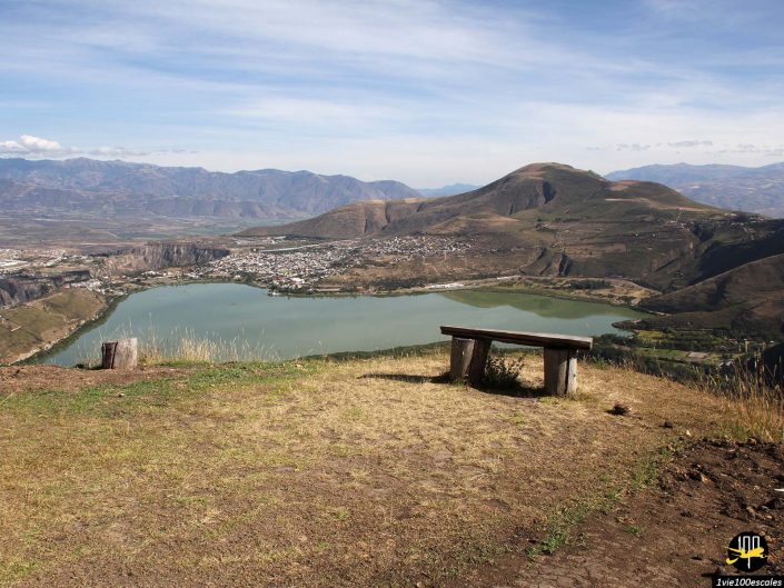 Un banc en bois au sommet d'une colline surplombe un lac serein entouré de montagnes et une petite ville dans la vallée en contrebas, sous le ciel bleu clair d'Ibarra en Équateur.