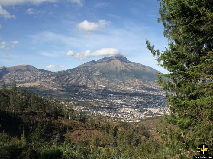 Une ville nichée dans une large vallée avec de nombreux bâtiments et entourée de collines verdoyantes et de forêts. En arrière-plan, une haute montagne au sommet couvert de nuages s'élève sous un ciel bleu à Ibarra en Équateur.