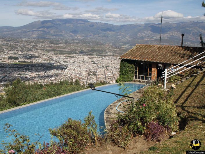 Piscine à débordement surplombant une ville avec des montagnes en arrière-plan à Ibarra en Équateur. Une petite maison en pierre avec un toit de tuiles jouxte la piscine, entourée de buissons floraux. Le ciel est partiellement nuageux.