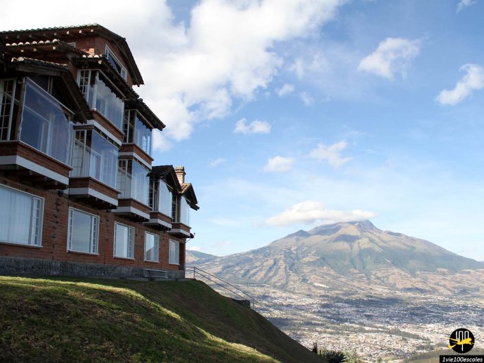Grande maison en brique dotée de multiples grandes fenêtres à flanc de colline avec vue sur un paysage urbain et des montagnes sous un ciel partiellement nuageux, à Ibarra en Équateur.