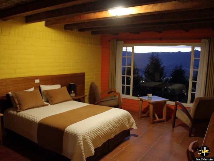 Une chambre douillette avec un lit double, deux chaises, un plafond en bois et une grande fenêtre offrant une vue sur le paysage montagneux au crépuscule, nichée dans le charmant lieu d'Ibarra en Équateur.