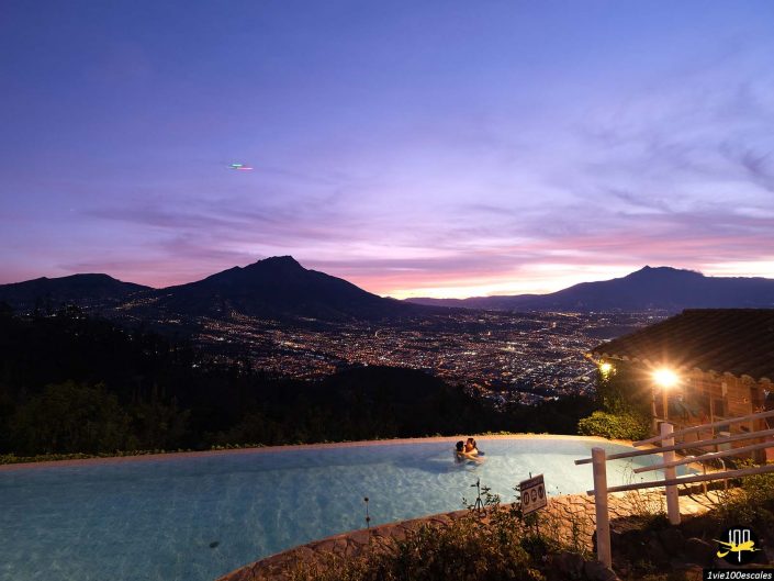 Une personne est assise au bord d’une piscine à débordement, surplombant un paysage urbain animé et des montagnes sous un ciel crépusculaire, à Ibarra en Équateur.