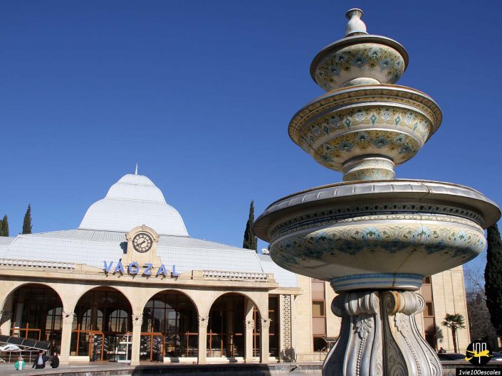 L'image montre un bâtiment en forme de dôme sur lequel est écrit « VAGZAL » et une grande fontaine décorative au premier plan, sur un ciel bleu clair, à Gandja en Azerbaïdjan.
