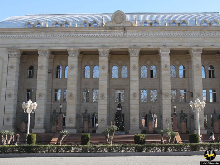Un grand bâtiment avec de grandes colonnes et une façade symétrique à Gandja en Azerbaïdjan présente un sapin de Noël à l'entrée. La structure possède de multiples fenêtres et éléments décoratifs, avec des lampes et des arbustes devant.