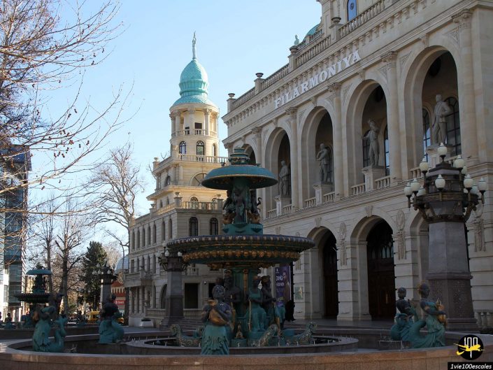 Une fontaine décorative ornée de statues de chérubins se trouve devant un grand bâtiment orné marqué "Filarmoniya" avec des détails architecturaux complexes et une tour en forme de dôme en arrière-plan, à Gandja en Azerbaïdjan.