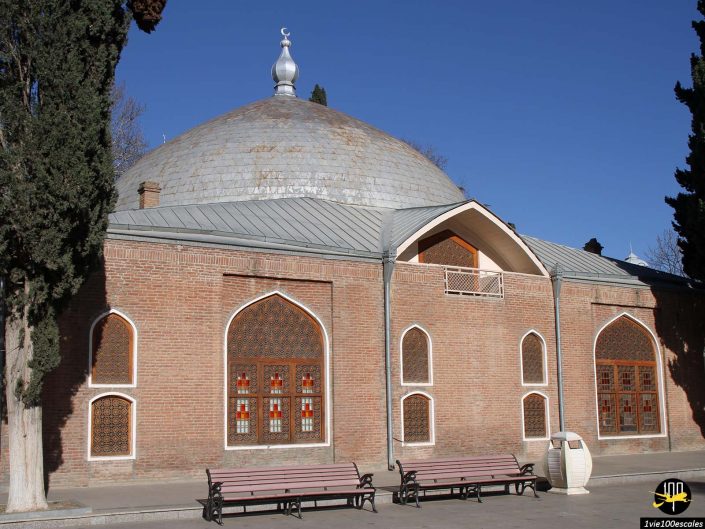 Un grand bâtiment en brique avec des fenêtres cintrées, un dôme argenté et des éléments architecturaux persans se dresse bien en vue à Gandja en Azerbaïdjan. Des bancs et des arbres entourent gracieusement la structure.