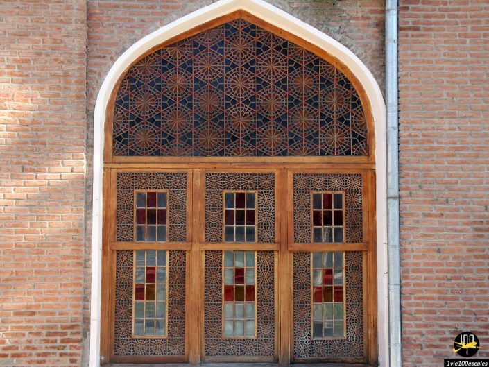Une porte en bois aux motifs géométriques complexes et aux petites vitres encastrées dans un mur de briques, à Gandja en Azerbaïdjan. La porte est surmontée d’un motif arqué comportant un treillis géométrique supplémentaire.