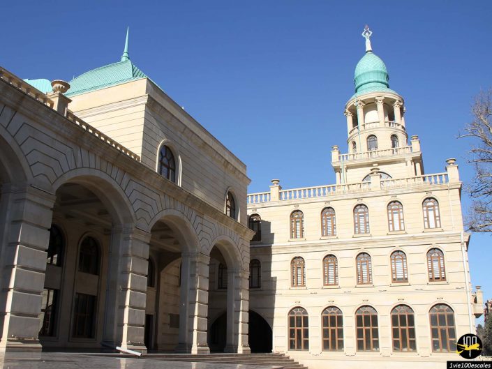 Un bâtiment en pierre beige avec des fenêtres cintrées et un dôme vert sous un ciel bleu clair se trouve à Gandja en Azerbaïdjan. La structure comporte une haute tour sur le côté droit ornée d'un dôme vert similaire.