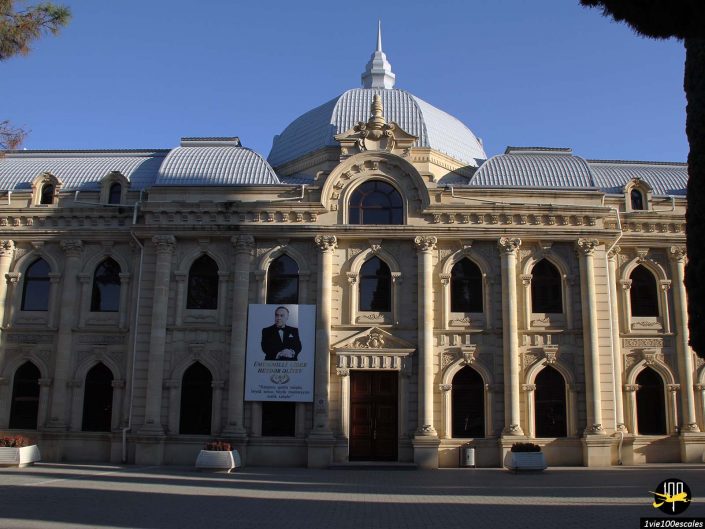 Un bâtiment historique avec des fenêtres cintrées et un toit en forme de dôme central se dresse sous un ciel clair à Gandja, en Azerbaïdjan, affichant une grande bannière verticale représentant une personne près de l'entrée.