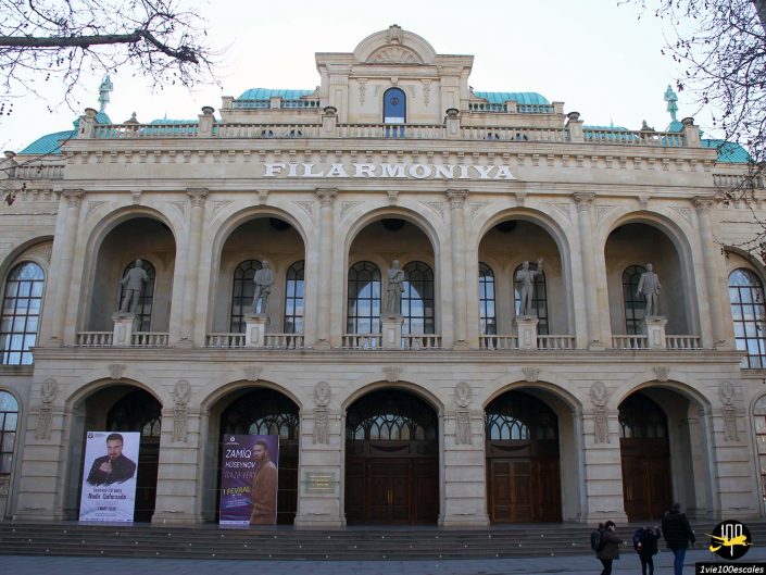 Façade du bâtiment Filarmoniya à Gandja en Azerbaïdjan avec fenêtres cintrées et statues dans des niches. De grandes banderoles sont accrochées de chaque côté de l’entrée et des personnes sont visibles près des portes.