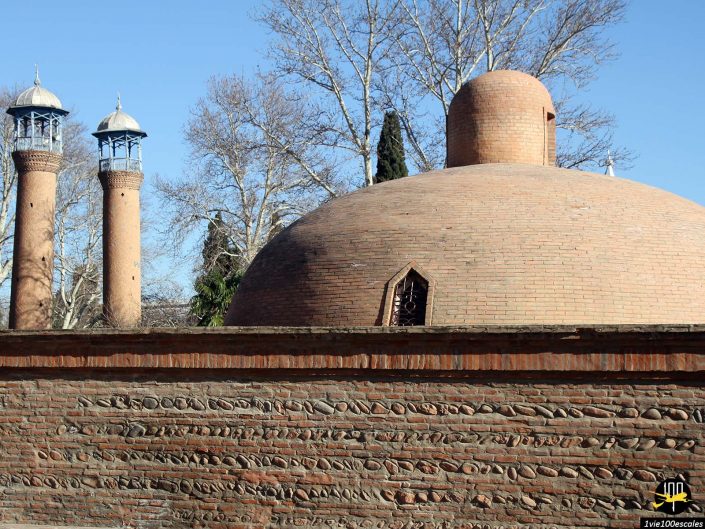 Une structure en brique avec un grand dôme et deux hautes tours en forme de minaret en arrière-plan, sur un ciel bleu clair et des branches d'arbres nues, à Gandja en Azerbaïdjan.