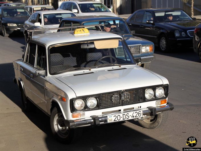 Un taxi blanc avec un panneau « TAXI » sur le dessus et la plaque d'immatriculation « 20-DS-426 » circule dans une rue urbaine très fréquentée à Gandja en Azerbaïdjan. D'autres véhicules sont visibles en arrière-plan.
