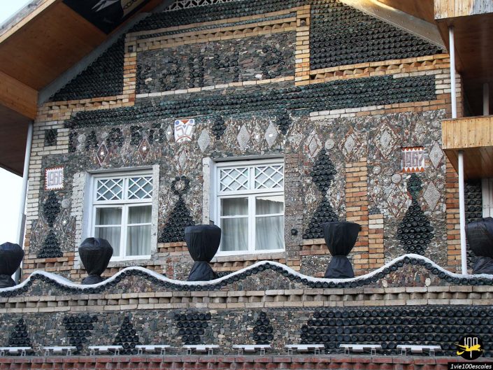 Une façade de maison à Gandja en Azerbaïdjan est décorée de bouteilles de couleurs variées et de pierres aux motifs géométriques. Les doubles fenêtres aux cadres blancs sont centrées et des sculptures enveloppées de tissu noir se trouvent sur le bord du balcon.