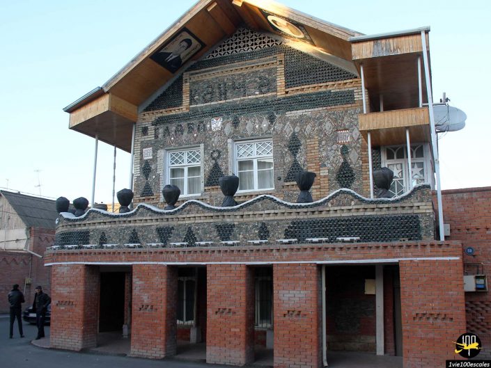 L'image montre un bâtiment de deux étages à Gandja en Azerbaïdjan avec une façade unique faite de bouteilles en verre et de briques. Le niveau supérieur présente des éléments décoratifs et des balcons, tandis que le niveau inférieur présente des entrées cintrées.
