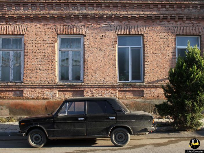 Une voiture vintage noire est garée dans la rue devant un vieux bâtiment en brique avec trois fenêtres et un petit arbre, à Gandja en Azerbaïdjan.