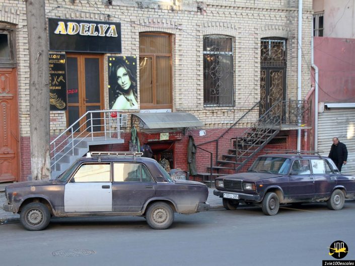 Deux modèles de voitures plus anciens sont garés dans une rue devant un immeuble avec une pancarte indiquant "Adelya" et une photo d'une femme, à Gandja en Azerbaïdjan. Une personne s’éloigne des lieux.