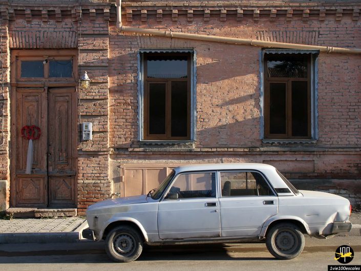 Une berline classique blanche est garée à Gandja en Azerbaïdjan devant un vieux bâtiment en brique avec deux fenêtres et une porte en bois ornée d'une couronne.
