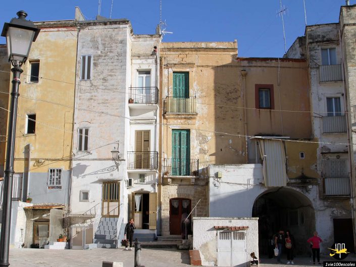 Une rangée de bâtiments connectés dans une zone urbaine à Bari en Italie, présentant divers détails architecturaux. Un groupe de personnes se tient dans le coin inférieur droit, près d’une arcade, et un lampadaire noir se trouve à gauche.