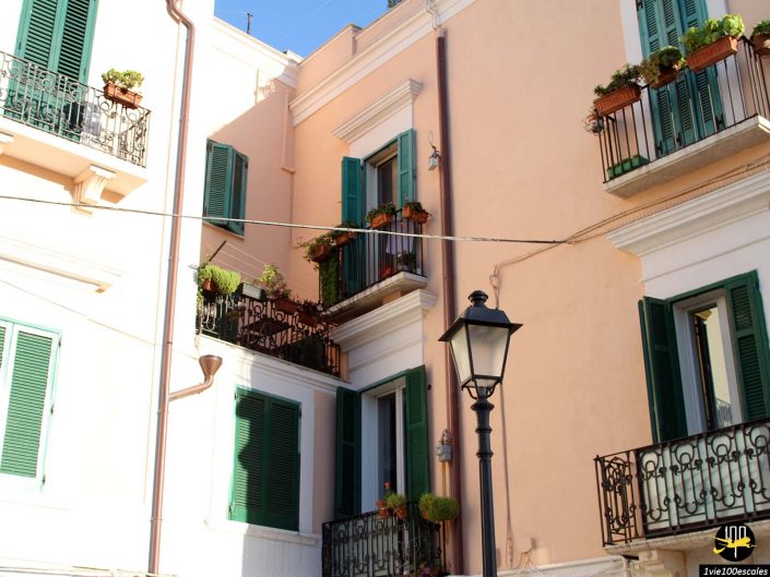 Façade d'un immeuble de style méditerranéen avec volets verts et balcons ornés de plantes en pot. Un réverbère est visible au premier plan, faisant allusion à une charmante scène à Bari en Italie.