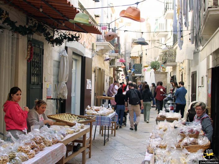 Un marché de rue étroit à Bari en Italie avec diverses tables installées vendant des produits de boulangerie. Les vendeurs et les acheteurs sont présents et des chapeaux colorés sont accrochés au-dessus de la rue.