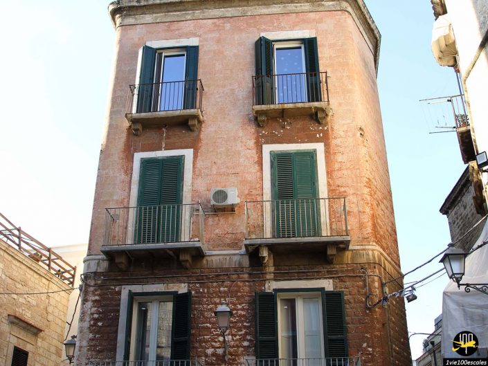 Un immeuble de trois étages avec une façade en brique rouge, des volets verts et des balcons en fer à Bari en Italie. Le bâtiment possède deux fenêtres à chaque étage et une unité de climatisation au deuxième étage.