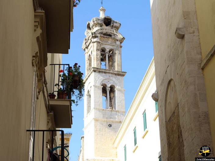 Vue sur une rue étroite menant à un vieux clocher en pierre avec une horloge à Bari en Italie. Des pots de fleurs ornent un balcon voisin. La lumière du jour met en valeur l’architecture.
