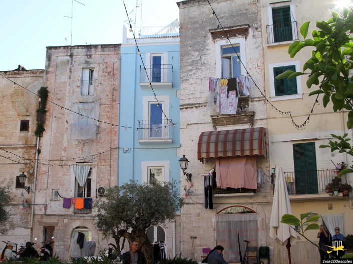 Scène de rue européenne historique à Bari en Italie, avec bâtiments altérés avec balcons, linge suspendu à l'extérieur et guirlandes lumineuses au-dessus. Les gens sont assis et se déplacent dans la zone.