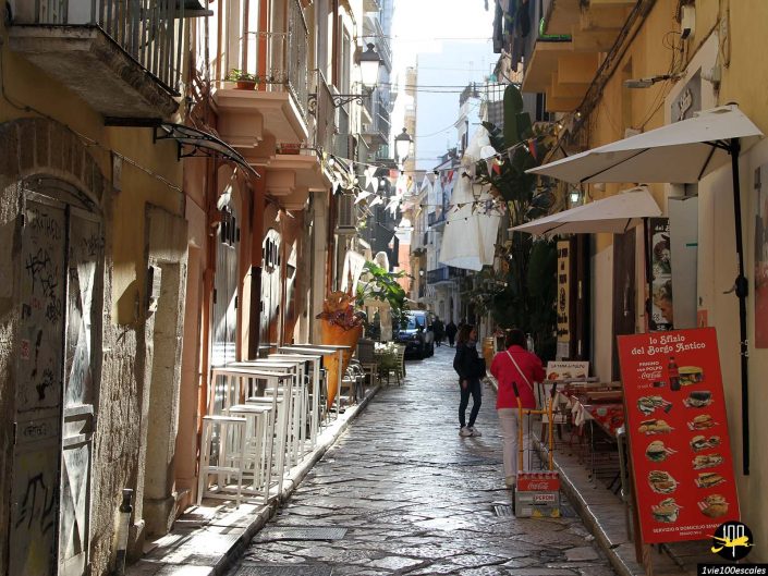 Une ruelle étroite à Bari en Italie, bordée d'immeubles, de sièges extérieurs et de luminaires suspendus, est peuplée de quelques piétons. La lumière du soleil illumine la scène, projetant des ombres sur le sol pavé de pierres.
