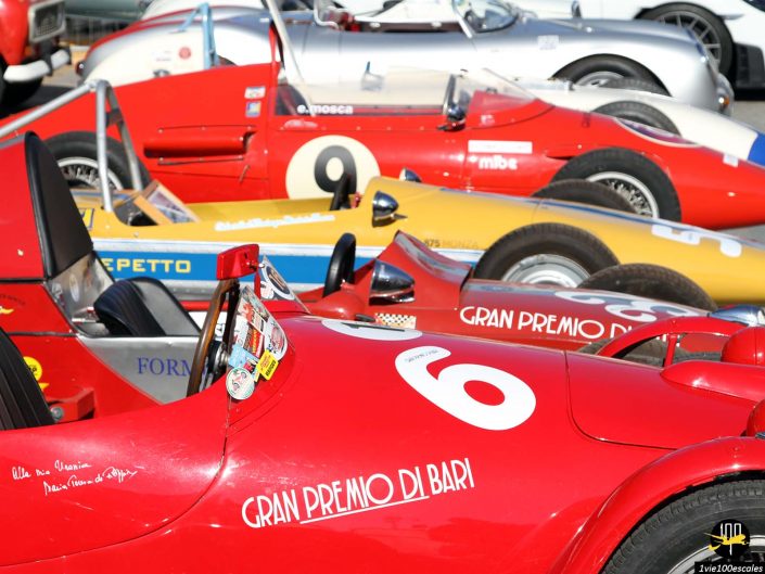 Ligne de voitures de course vintage avec des numéros bien visibles, pour la plupart en rouge, garées les unes à côté des autres lors d'un événement. La voiture avant porte le numéro « 6 » et est étiquetée « Gran Premio di Bari », capturant l'essence du sport automobile classique à Bari en Italie.