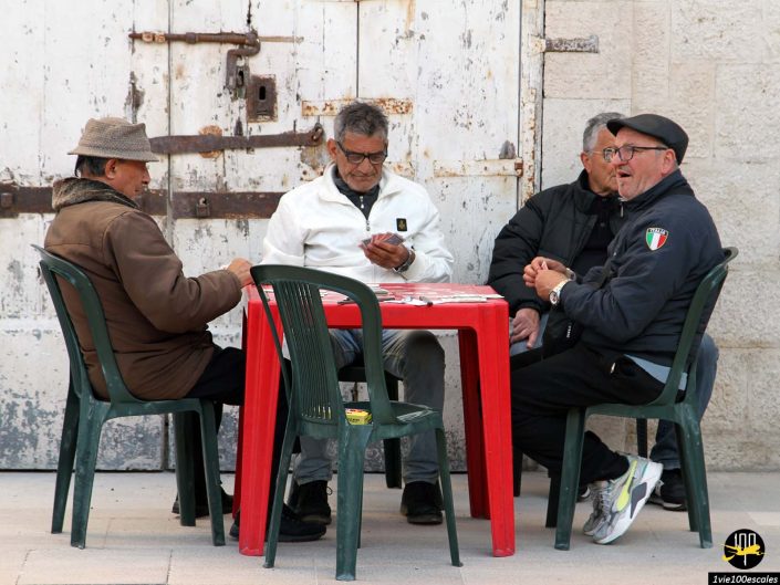 Quatre hommes sont assis autour d’une table en plastique rouge et jouent aux cartes à l’extérieur, près d’un mur blanc rustique. Vêtus de vêtements décontractés, ils semblent profondément engagés dans leur jeu, capturant une scène typique de Bari en Italie.
