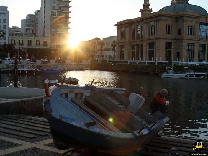 Un bateau altéré repose sur un quai au coucher du soleil, avec un bâtiment au bord de l'eau et plusieurs bateaux visibles en arrière-plan, à Bari en Italie.