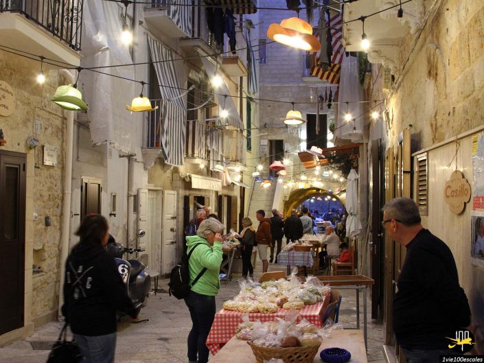Une rue étroite et pavée dans une petite ville de Bari en Italie avec des guirlandes lumineuses au-dessus. Les gens parcourent les marchandises sur les tables. Un vendeur de pâtisseries sert des clients à proximité d'un restaurant nommé Caffè Casino.