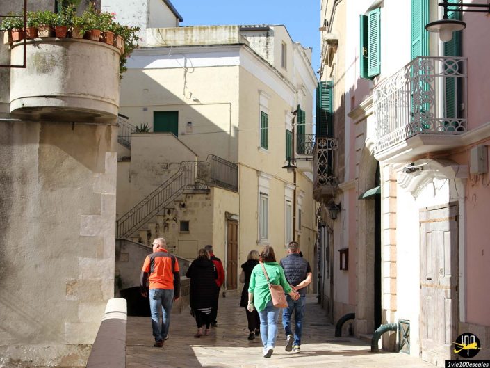 Un groupe de personnes marche dans une ruelle étroite et ensoleillée bordée de bâtiments anciens, certains avec balcons et plantes, à Matera en Italie.